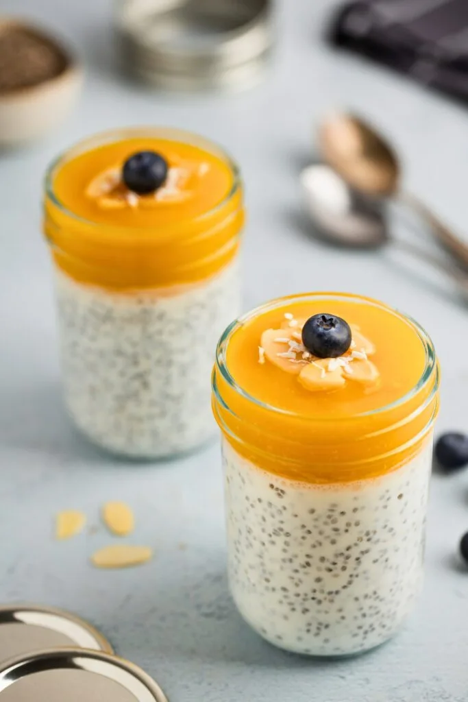 A jar of mango chia pudding topped with fresh blueberries, nuts, and shredded coconut